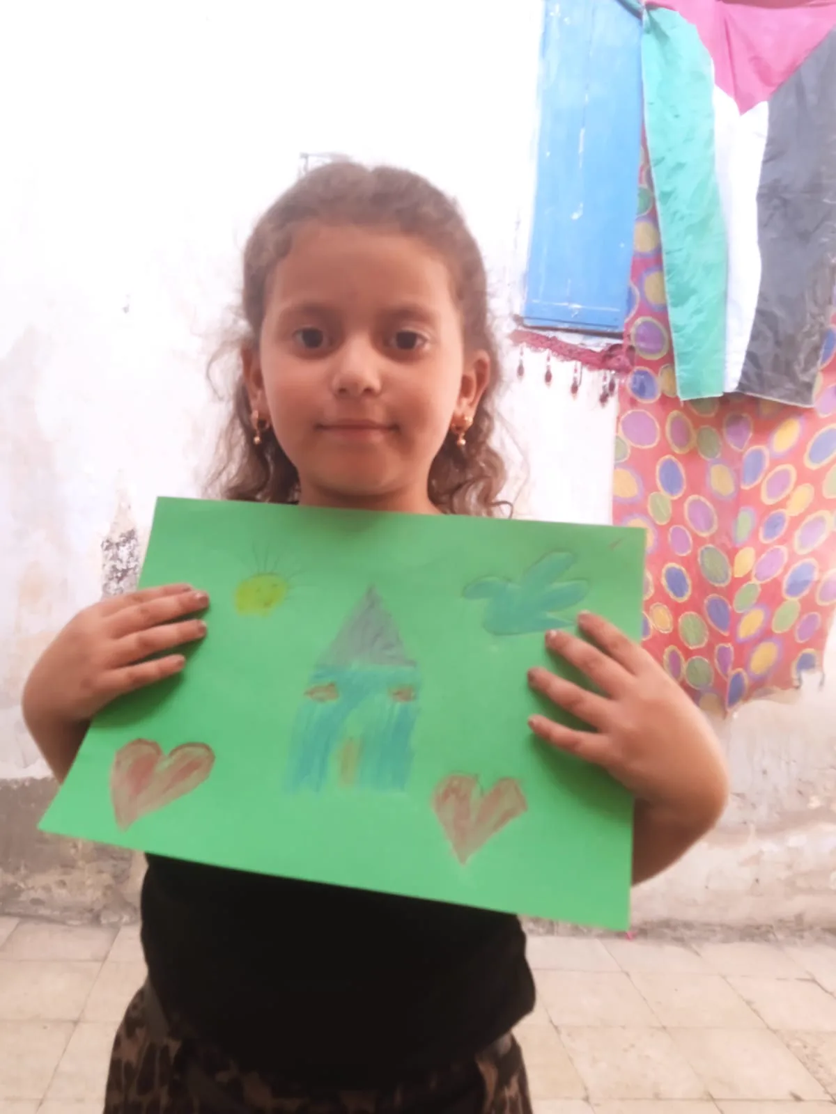 A young girl smiling softly as she holds up her drawing of a house, a sun, a bird and two hearts on green paper